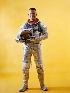 a man in an astronaut suit is holding a helmet and looking at the camera while standing against a yellow background