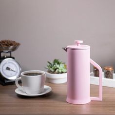 a white coffee pot sitting on top of a wooden table next to a cup of coffee