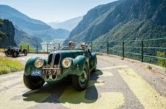 an old green car parked on the side of a mountain road next to a lush green valley