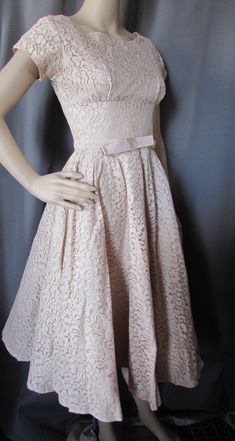 "Pretty lace party dress in a buttercream tone of beige.  Typical of late'50s, early '60s design, it has a tightly fitted empire bodice and full skirt. The waist is set slightly lower than the natural waist and is highlighted with a self fabric belt with 6\" bow at front. It has 4 1/2\" cap sleeves. The back plunges 5 1/2\" to 15\" metal zipper. The under bust seam is 9 1/2\" from throat. The dress is lined. No maker label Size: no size label--35\" bust; 27\" at the natural waist; 34\" at slight Beige A-line Lace Dress, Fitted Beige A-line Lace Dress, Beige Fitted A-line Lace Dress, Beige A-line Lace Wedding Dress, Cream A-line Lace Dress, Elegant Cream Vintage Dress For Garden Party, Cream Lace Evening Dress Feminine Style, Feminine Cream Lace Evening Dress, Cream Lace Dress For Evening