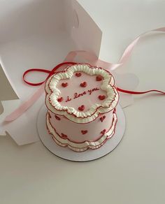 a pink cake sitting on top of a table next to a white box with a red ribbon