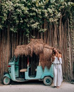 a woman standing next to a small car with a straw hut on the roof and palm trees in the background