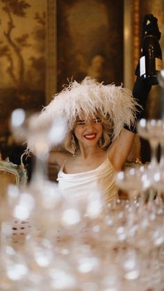 a woman in a white dress and feather hat sitting at a table with wine glasses