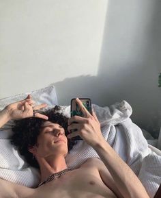 a shirtless man laying in bed holding up his cell phone to take a selfie