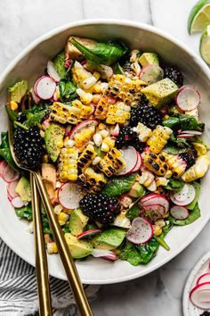 a white bowl filled with mixed vegetables and grilled corn on the cob next to two gold serving utensils