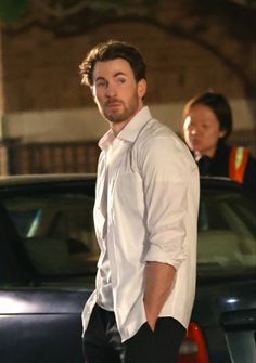 a man in white shirt and black pants standing next to a car with his hand on his hip