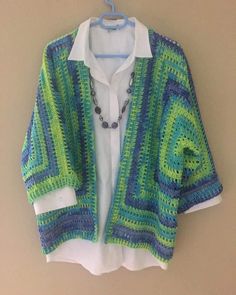 a green and blue crocheted cardigan hanging on a hanger next to a white shirt