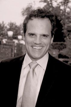 a black and white photo of a man in a suit smiling at the camera with trees in the background