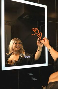 a woman is looking at herself in the mirror and writing on her face with orange marker