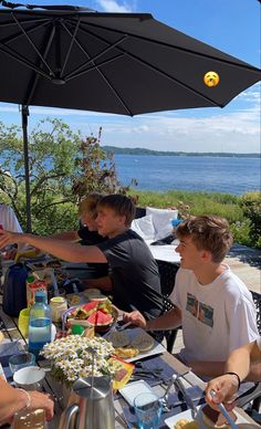 people sitting at an outdoor table with food and drinks on it, near the water