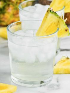 two glasses filled with ice and some pineapples in the background on a table