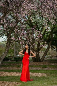 a woman in a red dress standing under a tree with pink flowers on it's branches