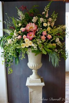 a white vase filled with lots of flowers on top of a stone pillar next to a wall