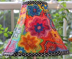 a colorful lamp shade is hanging on a wooden stand in front of some flowers and greenery