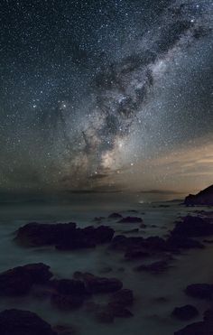 the night sky is filled with stars and clouds above rocks in the ocean, as seen from an island