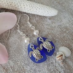 two blue sea turtle earrings with pearls and seashells on the sand next to a starfish shell