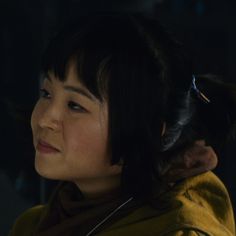 a woman with dark hair wearing a yellow jacket and looking off to the side, in a darkened room