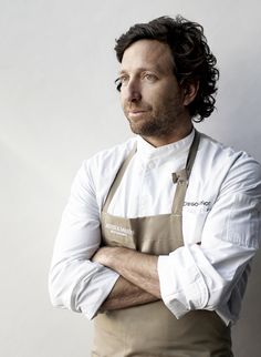 a man with his arms crossed standing in front of a white wall wearing an apron