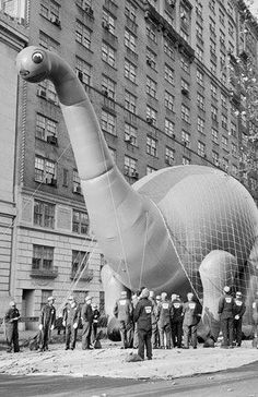 an inflatable dinosaur balloon is being flown by men on the street while others watch