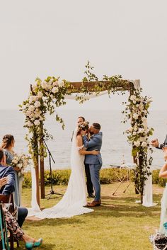 a bride and groom are kissing under an arch decorated with flowers on the lawn by the ocean