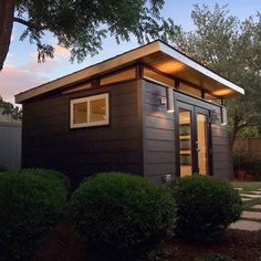 the tiny house is located in front of a tree and bushes, with its lights on