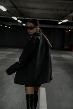 a woman standing in an empty parking lot wearing knee high boots and a black coat