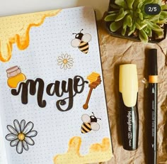 a notebook with the word may written on it next to some pens and a plant