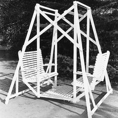 two white chairs sitting next to each other on a dirt ground near trees and bushes