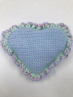 a crocheted blue heart shaped pillow on a white surface with green and pink trim