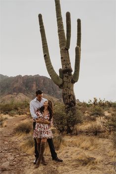 a man and woman standing in front of a large saguado cactus with their arms around each other