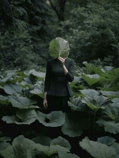 a woman standing in the middle of a lush green forest holding a leaf over her face