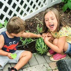 two young children are playing with watermelon in the garden, one is biting into the other's hand