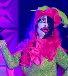 a woman in a clown mask and green dress on stage with her hands out to the side