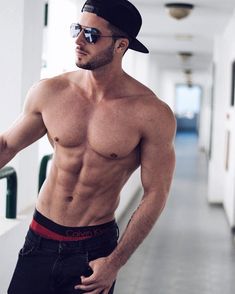 a shirtless man wearing sunglasses and a hat is standing in a hallway with his hands on his hips