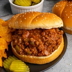 two sloppy joes on a plate with pickles and chips next to them,