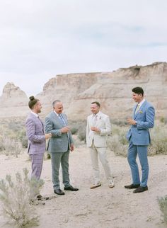 three men in suits talking to each other on a dirt road with mountains in the background