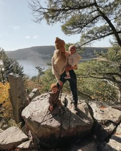 a woman holding a baby standing on top of a large rock next to a tree