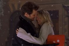 a man and woman embracing each other in front of a fire place with snow falling on the ground