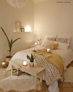 a white bed sitting next to a wooden table with candles on it and a plant in the corner