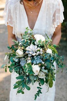 a woman holding a bouquet of flowers and greenery in her hands while wearing a white dress