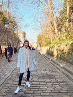 a woman standing in the middle of a cobblestone road wearing a blue coat