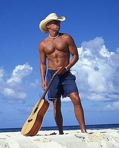 a man standing on the beach holding a guitar and wearing a cowboy's hat