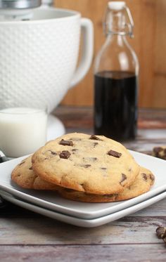 two chocolate chip cookies on a white plate next to a glass of milk and coffee