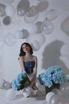 a woman sitting on the ground surrounded by blue and white flowers with balloons in the background