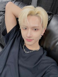 a young man with blonde hair sitting on a black leather chair and wearing a chain around his neck