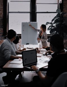 a group of people sitting around a table in front of a whiteboard with a woman pointing at it