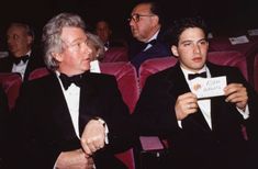 two men in tuxedos sitting next to each other at an event, one holding a sign