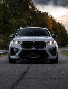 the front end of a white bmw suv driving down a road with trees in the background