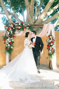 a bride and groom kissing under an archway decorated with red, white and pink flowers