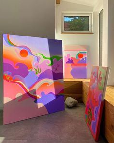 three colorful paintings sitting on top of a floor next to each other in front of a window